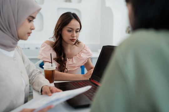 Three young coworkers gather around laptop in bright modern office, with focused woman in pink blouse studying details while others review documents and share ideas beside iced drink.