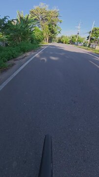 Vertical POV footage of a cyclist casting a long shadow on a quiet rural road, with trees, houses and warm sunlight creating a peaceful countryside scene.