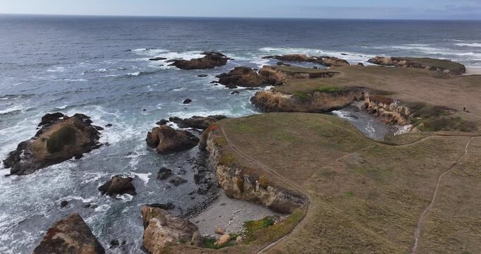 Aerial view of the rugged coastline and rocky cliffs at Glass Beach with crashing waves and grassy trails in Fort Bragg, California, United States.