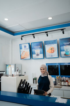 Portrait of cheerful female barista wearing hijab at cafe counter with tablet, coffee cups, drink equipment, and blue accented walls, showing friendly customer service and small business atmosphere