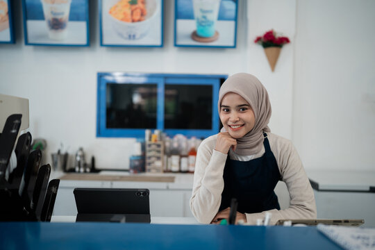 Portrait of smiling female shop worker in hijab standing behind modern counter with tablet in bright store interior, blue framed windows, product displays, and clean customer service setting.