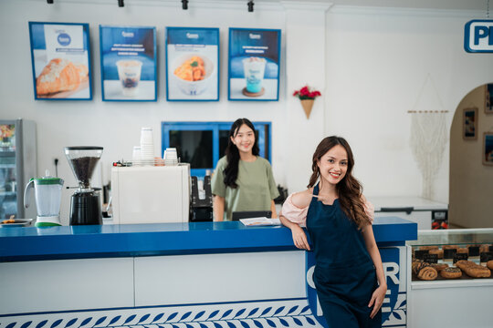 Smiling woman in apron leans on blue counter inside bright bakery cafe, with coffee grinder, drink area, pastry display, menu boards, and another employee in background.