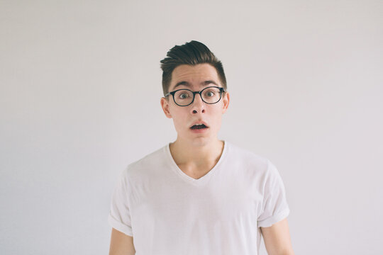 Omg No way Very shocked young man with glasses is looking at the camera. Student nerd is wearing in a white t-shirt, standing isolated on a white background