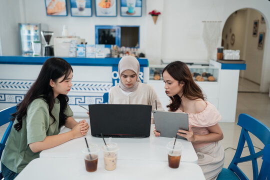 Three women in bright cafe work together at table, reviewing on laptop and tablet during focused business discussion with iced drinks and modern seating nearby.