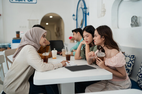 Four young adults sit face to face in bright cafe interior, talking over iced drinks, papers, and tablet on white table, creating relaxed social meeting scene with modern minimal decor.