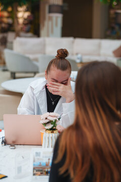 Stressed young woman covering face crying during therapy session or difficult conversation in office. Mental health crisis, burnout and depression concept