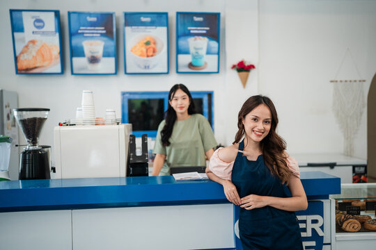 Smiling woman in navy apron leans on bright blue counter inside modern dessert shop, with coworker behind coffee machine, menu posters on wall, white interior, and friendly retail service mood.