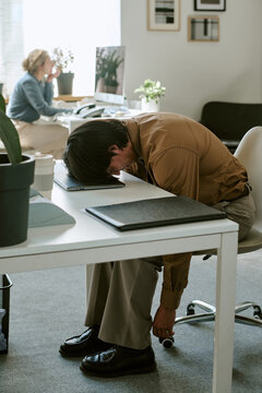Vertical shot of exhausted young adult businessman sleeping on laptop at workplace desk. Scene depicting corporate burnout, suitable for mental wellbeing and work stress campaigns