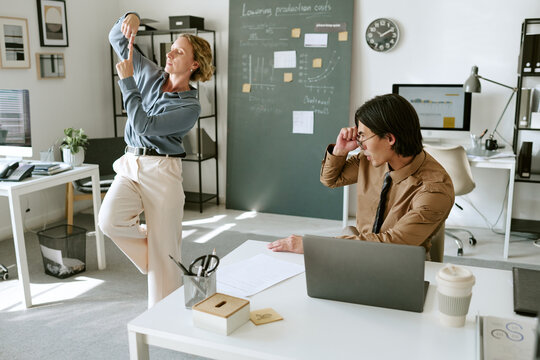 Middle aged Caucasian woman dancing while adult man sitting at laptop watching in modern office. Concept of workplace stress relief and corporate culture for human resources advertising