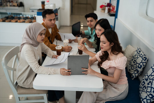 Five young adults gather around table in bright cafe interior, reviewing documents on tablet and laptop while talking over iced drinks and notes in relaxed collaborative workspace.