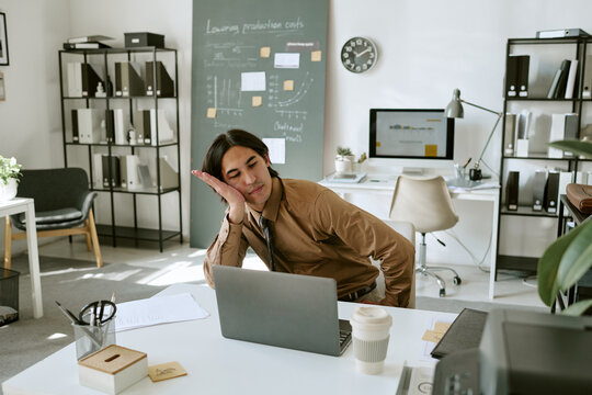 Tired young adult businessman sitting at desk looking at laptop screen experiencing workplace burnout. Image suitable for business stress and corporate mental health campaigns