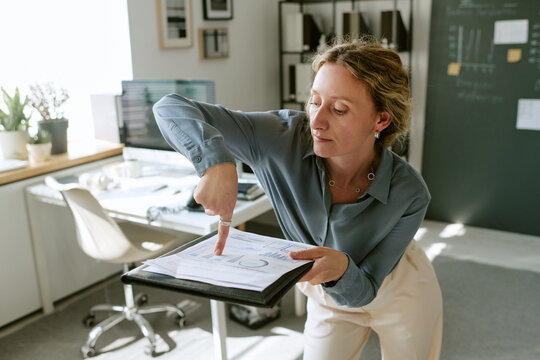 Middle aged Caucasian woman pointing at paper chart in folder while analyzing statistics in office. Image suitable for business analysis, corporate reporting, financial strategy campaigns