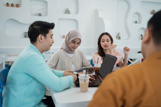 Small group of young adults seated around laptop in bright modern cafe, reviewing notes and discussing project with friendly, focused expressions and casual drinks.