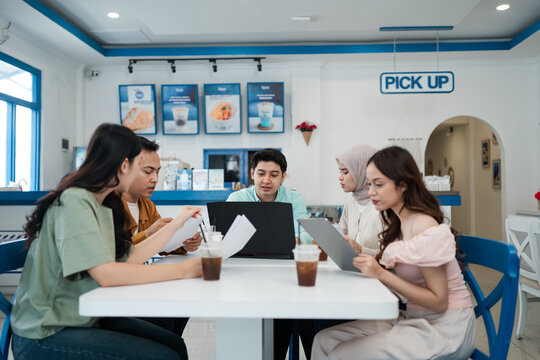 Five young adults sit together at bright cafe table, reviewing documents and laptop while sharing drinks in modern coffee shop, suggesting teamwork, planning, study, and informal business discussion.