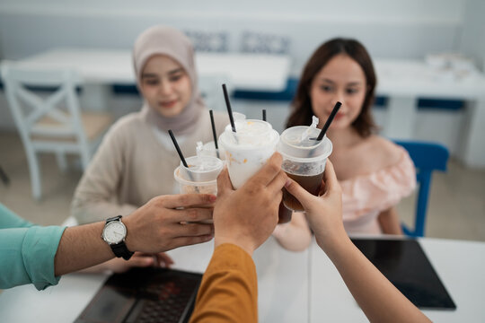 Hands clink plastic cups filled with iced coffee and creamy drinks on modern cafe table, with two softly blurred women seated behind in bright daylight, creating relaxed toast between friends.