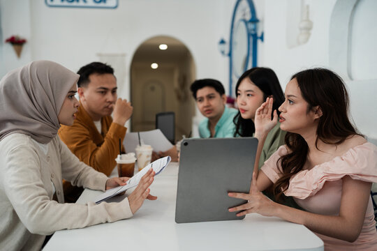 Four young adults collaborate around laptop with papers and coffee drinks in bright cafe, using focused hand gestures and friendly conversation during shared planning.