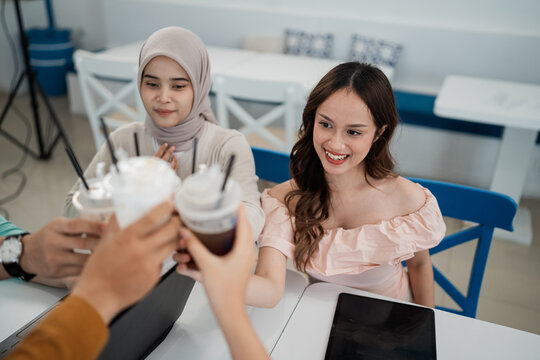 Smiling young women sit together at bright cafe table while raising iced drinks in friendly toast, with tablet on tabletop, soft daylight, blue chairs, and relaxed social atmosphere.