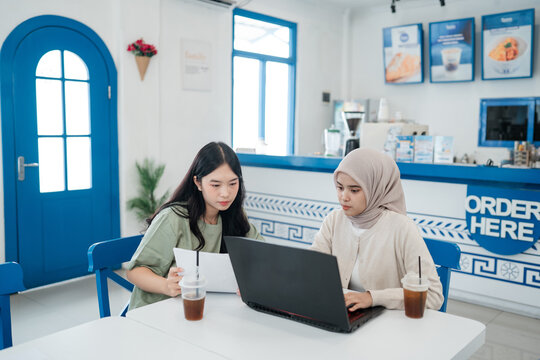 Two women at table in sunlit blue cafe review notes on laptop while sharing takeaway coffee drinks, creating focused teamwork mood for study, remote work, and planning.