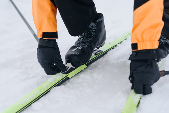 young active skier fastens ski fastenings on legs before skiing in snowy field, sportive caucasian man in orange and black clothes, ski poles in hands, cropped, cloudy winter day, healthy lifestyle