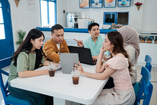 Five young adults sit around white table in bright cafe workspace, discussing project on laptop and tablet with casual drinks nearby, focused teamwork and brainstorming.