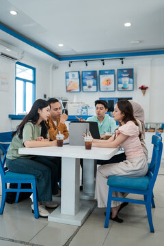 Four young adults sit together in bright cafe, sharing iced drinks while discussing around laptop and tablet. Modern blue and white interior, casual teamwork, friendship, and conversation.
