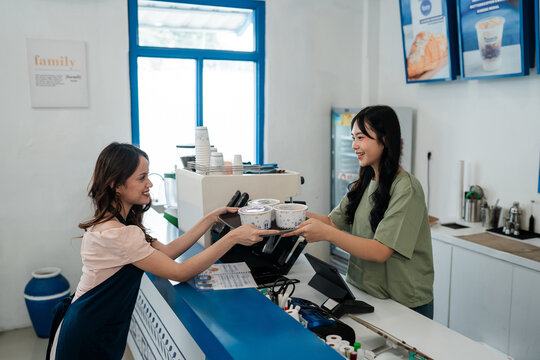 Smiling women hand dessert cups to customers at bright cafe counter, showing teamwork, hospitality, and clean modern food shop interior with menu posters and counter equipment.
