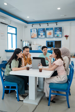 Five young adults sit together at bright modern cafe table, sharing ideas around laptop and tablet with drinks nearby, relaxed teamwork, study collaboration, and casual business meeting atmosphere.