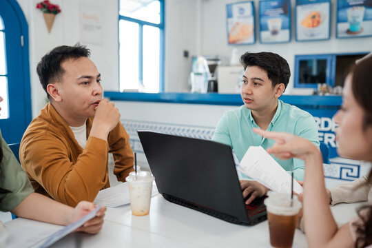 Group of young professionals seated around laptop at bright cafe table, reviewing documents and notes while discussing strategy, sharing advice, and coordinating next steps for project.