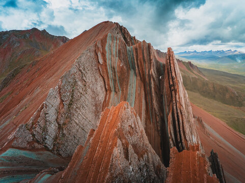 Aerial view of the striking jagged peaks of Pallay Punchu Rainbow Mountain with its distinct red and turquoise mineral layers under a dramatic cloudy sky in Layo, Cuzco, Peru.