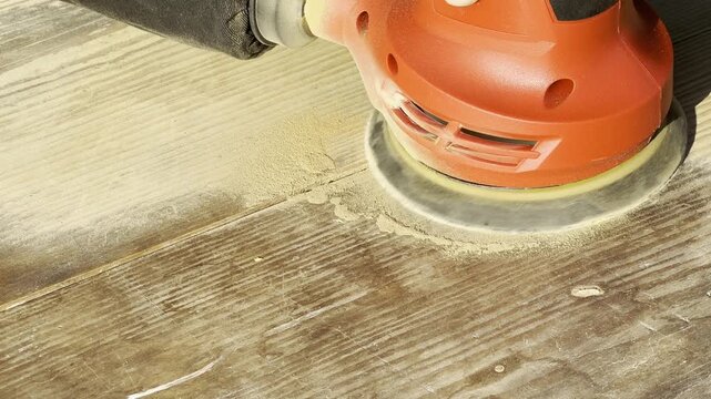 Close-up of orbital sander working on old wooden table, rotating abrasive disc removing worn surface layer and producing fine wood dust during DIY furniture restoration at home, power tool in operatio