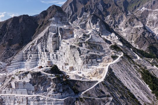 Aerial view of the famous white marble quarries in the Apuan Alps, showing terraced excavation sites, winding access roads, and rugged mountain terrain in Carrara, Tuscany, Italy.