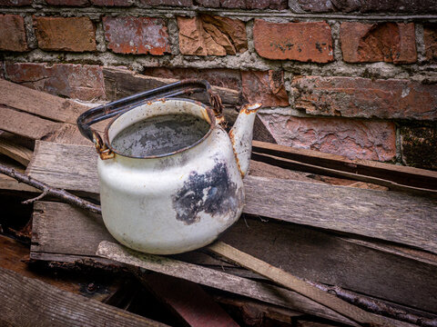 A vintage, weathered white enameled kettle with noticeable rust spots, soot, and missing enamel sections resting on a pile of old wooden planks and laths. In the background, a rustic, textured red 