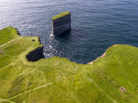 Aerial view of Dun Briste Sea Stack rising from the Atlantic Ocean near grassy cliffs with the historic EIRE 64 sign visible in County Mayo, Ireland.