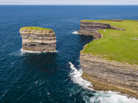 Aerial view of Dun Briste sea stack rising from the Atlantic Ocean near the dramatic cliffs of Downpatrick Head with lush green grass and layered rock formations, County Mayo, Ireland.