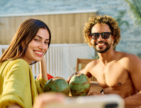 Two people enjoy coconuts while sitting in water at a pool area and using a smartphone camera  taking a selfie photo in a warm location during summer