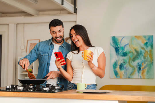 A happy couple cooks side by side at a kitchen island, one holding a red phone while the other stirs a frying pan and wears a white top. Nearby a mug sits on the counter as they smile and share a
