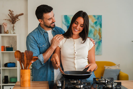 A warm scene shows a couple enjoying food prep at a stove, one stirs a skillet while the other smiles and assists. Wooden utensils stand in a holder, emphasizing teamwork and shared cooking