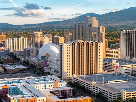 Aerial shot of the urban skyline in Reno, Nevada, displaying casino resort high rises, the Silver Legacy dome, streets, and distant mountain ranges in warm evening light.