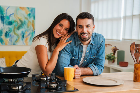 A cheerful couple shares a warm moment in a sunlit kitchen while making coffee. Hands meet, a yellow mug rests between them, and a gas stove and utensils sit nearby for a cozy culinary scene