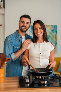A happy couple cooks together at a wooden counter, one holds a spatula while the other rests a hand on the shoulder, a pan heats on the stove, and warm smiles convey warmth and teamwork in the moment.