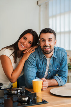Two smiling partners lean toward each other over a wooden table, hands near a yellow mug. The image captures warm companionship, casual conversation, and a comfortable morning routine in a kitchen now