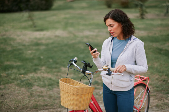 Woman standing next to her bicycle, holding a smartphone and looking at the screen with a focused expression, navigating a map or interacting with an app in a park