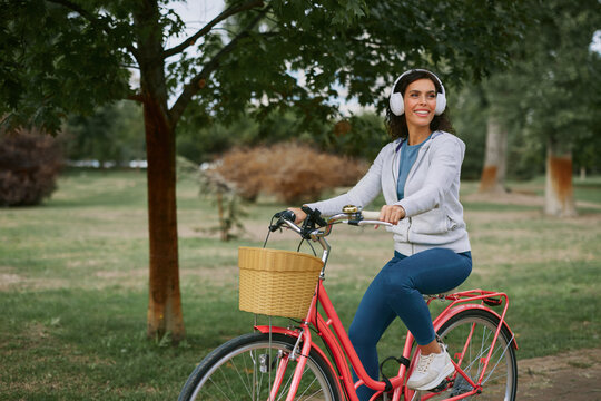 Woman riding bicycle with a front basket and white headphones, smiling and looking away while enjoying a sunny park ride, fresh air, leisure and active healthy lifestyle