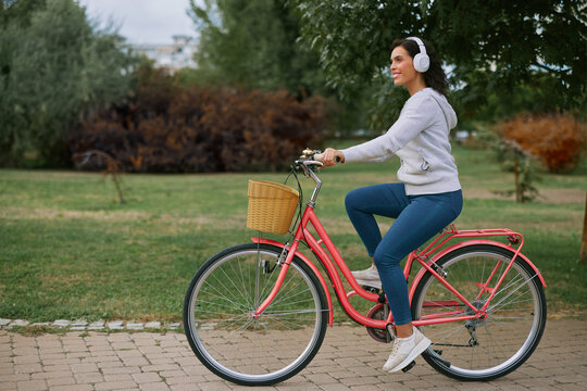 Young woman enjoying an active outdoor lifestyle, cycling on a pink bicycle through a park while listening to music with headphones, embracing wellness and leisure