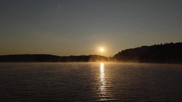 Cinematic drone shot flying low over a calm Muskoka lake through floating mist, then gradually ascending toward a glowing sunset above dark forested shores in Ontario, Canada.