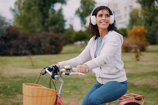 Woman enjoying a leisurely bicycle ride in a city park, wearing wireless headphones and smiling, embodying happiness, freedom, and an active lifestyle