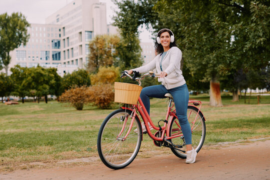 Happy woman enjoying urban leisure, riding her bicycle in a city park while listening to music with headphones, embracing active outdoor lifestyle and freedom