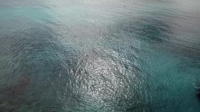 A slow-moving aerial drone shot looking straight down at the rippling blue and turquoise sea surface, with bright sunlight reflecting and sparkling across the gentle waves.