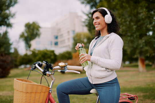 Woman pausing during a bike ride in the park, holding a water bottle and wearing headphones, smiling and looking away while finding refreshment and relaxation outdoors