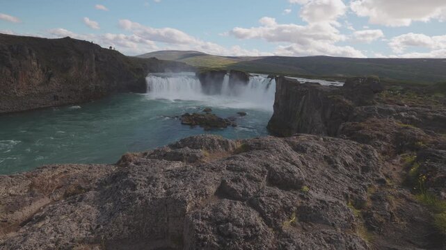 Godafoss waterfall pours into a turquoise river between dark volcanic rock formations in northern Iceland, establishing static wide overview